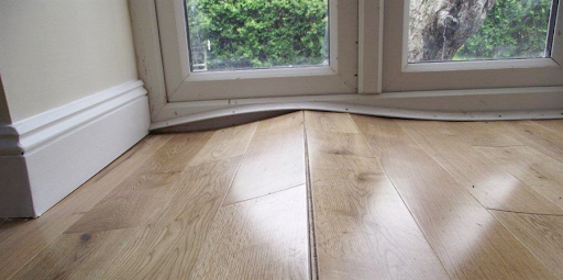Warped wooden floorboards lifting near a window and skirting board.